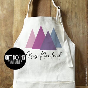 May include: White apron with a design of three pink, purple, and blue triangles. The text "Mrs. Nordack" is printed in black cursive lettering.