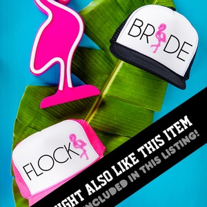May include: Two pink and white trucker hats with flamingo designs. One hat says "Flock" and the other says "Bride". The hats are on a green leaf with a pink flamingo figurine.