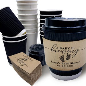 May include: An assortment of disposable coffee cups and lids. The image includes black corrugated cups, white cups, and a cup with a brown sleeve that reads "A Baby is Brewing" with a baby footprint design. The date 08.26.2026 is also printed.