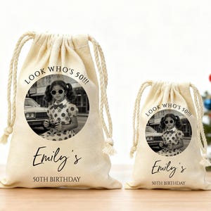 May include: Two beige drawstring bags with a black and white photo of a young girl wearing sunglasses. The bags have the text "LOOK WHO'S 50!!!", "Emily's", and "50TH BIRTHDAY". One bag is larger than the other.