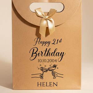 Pode incluir: Uma sacola de presente de papel castanho com al&ccedil;a e la&ccedil;o dourado. A sacola tem o texto "Happy 21st Birthday" e a data 10.10.2004, junto com uma ilustra&ccedil;&atilde;o de duas m&atilde;os brindando com ta&ccedil;as de coquetel. O nome "HELEN" est&aacute; impresso na parte inferior.