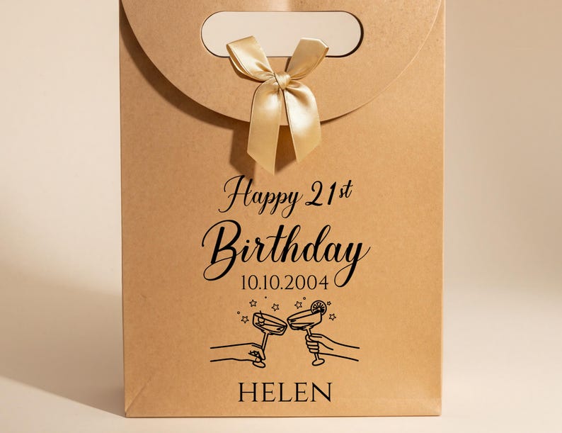Pode incluir: Uma sacola de presente de papel castanho com al&ccedil;a e la&ccedil;o dourado. A sacola tem o texto "Happy 21st Birthday" e a data 10.10.2004, junto com uma ilustra&ccedil;&atilde;o de duas m&atilde;os brindando com ta&ccedil;as de coquetel. O nome "HELEN" est&aacute; impresso na parte inferior.