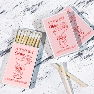May include: Pink matchboxes with the text "A Tini Bit Older" and "Lauren's 30th Birthday" in red. The design includes a cocktail glass with a lemon slice and a bow. The matches are visible inside one box.