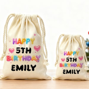 Op de afbeelding: Twee beige geschenkzakken met trekkoord met de tekst "HAPPY 5TH BIRTHDAY EMILY" in kleurrijke bubbelletters. De tassen hebben roze harten en een cadeau-doos afbeelding. De tassen staan op een houten oppervlak.