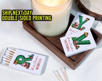 May include: A lit candle sits on a wooden tray with three matchboxes. The matchboxes are white with a green letter, Christmas illustrations, and personalized names. The text "Merry Christmas" is also visible.
