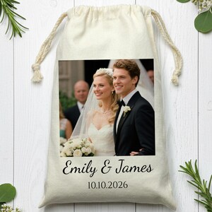 May include: A cream-colored drawstring bag featuring a wedding photo of a smiling couple. The bag is printed with the names "Emily & James" and the date "10.10.2026" in black script. The bag is decorated with greenery.