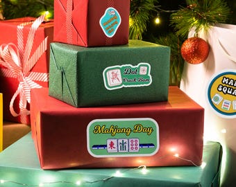 May include: Wrapped gift boxes in red, green, and teal with Mahjong-themed stickers. The stickers feature text and Mahjong tile illustrations. A Christmas tree and ornaments are in the background.