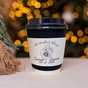 May include: A black and white paper coffee cup with a black lid. The cup has a white band with the text "the perfect blend" and a line drawing of a couple. The names "Daniel + Lauren" and the date "03.21.2026" are also printed on the band.
