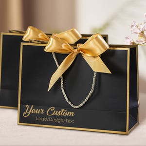 Op de afbeelding: Twee zwarte cadeauzakken met gouden randen en handvatten. Elke tas heeft een gouden satijnen strik en de tekst "Your Custom Logo/Design/Text" in gouden script. De tassen staan op een licht oppervlak.