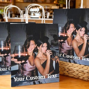 Op de afbeelding: Cadeautassen met witte handvatten met een zwart-wit foto van twee vrouwen die wijnglazen vasthouden. De tassen hebben de tekst "Your Custom Text" in het wit. De tassen staan op een houten tafel.