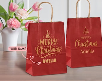 Puede incluir: Dos bolsas de regalo de papel rojo con asas marrones. Las bolsas tienen texto dorado que dice "Merry Christmas" y un nombre. Una bolsa tiene un diseño de árbol de Navidad. Un jarrón con flores rosas está en el fondo.