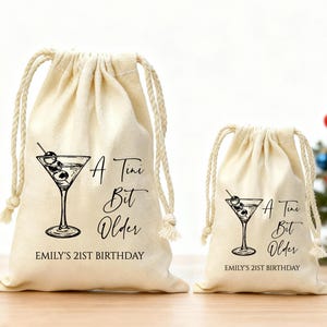 May include: Two beige drawstring bags with a martini glass design and the text "A Tiny Bit Older" and "Emily's 21st Birthday" in black. The bags have a rope drawstring closure.