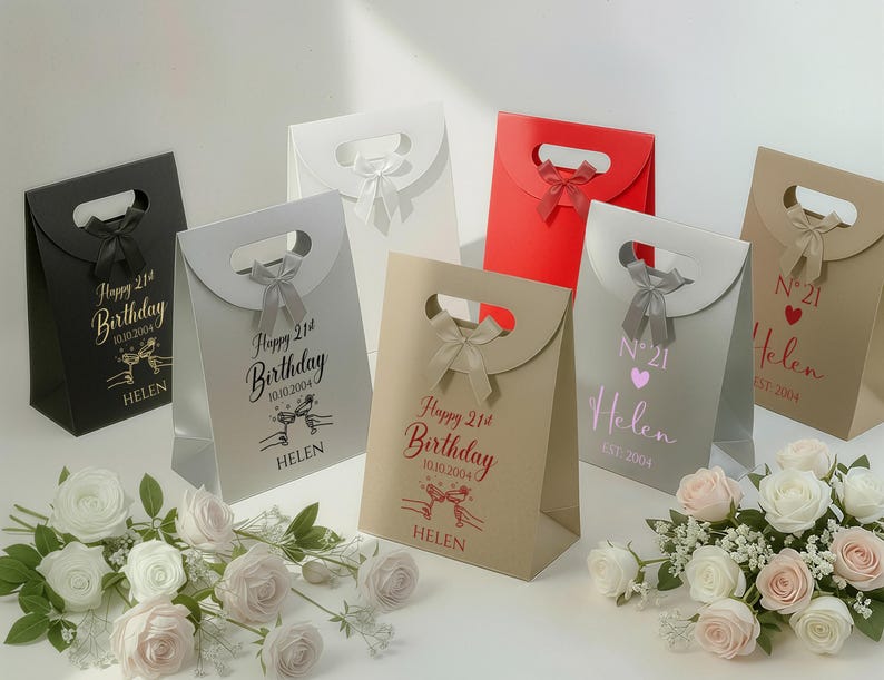 Pode incluir: Uma cole&ccedil;&atilde;o de sacolas de presente em v&aacute;rias cores, incluindo preto, branco, vermelho e bege. Cada sacola apresenta uma al&ccedil;a, um la&ccedil;o e texto personalizado, como "Happy 21st Birthday" e um nome. Arranjos florais em primeiro plano.