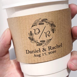 以下が含まれることがあります： 白い紙のコーヒーカップと茶色の段ボールスリーブ。スリーブには、イニシャル「D/R」と「Daniel & Rachel」の名前、日付「2026年8月17日」が記された黒い花のリースデザインが施されています。