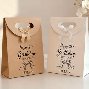Pode incluir: Duas sacolas de presente, uma bege e outra branca, com al&ccedil;a e la&ccedil;o. As sacolas t&ecirc;m a inscri&ccedil;&atilde;o "Happy 21st Birthday", a data, o nome "Helen" e um desenho de duas m&atilde;os brindando.