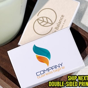 May include: White business cards with a teal, orange, and blue logo, and the text "COMPANY business tagline". A wooden tray holds the cards and a beige card with a leaf logo. The text "SHIP NEXT DAY DOUBLE-SIDED PRINTING" is also visible.