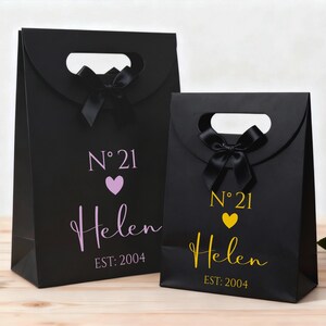 Pode incluir: Duas sacolas de presente pretas com al&ccedil;as recortadas e la&ccedil;os pretos. A sacola maior tem o texto "N&deg; 21" acima de um cora&ccedil;&atilde;o roxo e o nome "Helen" em it&aacute;lico, com "EST: 2004" abaixo. A sacola menor tem o mesmo design em amarelo.