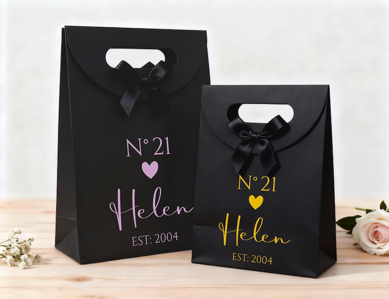 Pode incluir: Duas sacolas de presente pretas com al&ccedil;as recortadas e la&ccedil;os pretos. A sacola maior tem o texto "N&deg; 21" acima de um cora&ccedil;&atilde;o roxo e o nome "Helen" em it&aacute;lico, com "EST: 2004" abaixo. A sacola menor tem o mesmo design em amarelo.