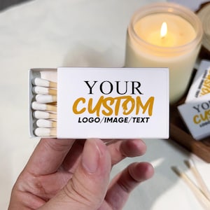 May include: A white matchbox with the text "YOUR CUSTOM LOGO/IMAGE/TEXT" in black and gold. The matchbox is open, revealing white-tipped matches. A lit candle in a glass jar and additional matchboxes are in the background.