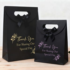 May include: Two black gift bags with a handle and a black bow. The bags feature the text "Thank You For Sharing Our Special Day" with an olive branch design. One bag has gold text, the other pink.