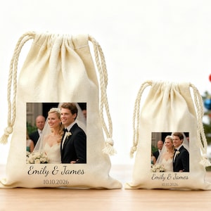 Puede incluir: Dos bolsas beige con cordón, cada una con una foto de boda de una pareja sonriente. Las bolsas tienen impreso el texto "Emily & James" y la fecha "10.10.2026". Las bolsas están sobre una superficie de madera.