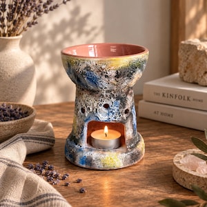 Pode incluir: Um queimador de óleo de cerâmica com um esmalte texturizado multicolorido em tons de azul, amarelo e preto. O topo apresenta uma bacia lisa de cor rosa claro, enquanto a base tem uma ampla abertura arqueada contendo uma pequena vela branca acesa.