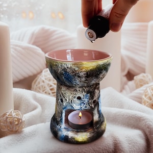 Könnte beinhalten: Ein Keramik-Diffusor für ätherische Öle mit rosa Innenraum und mehrfarbigem Äußeren. Der Diffusor hat eine brennende Teelichtkerze im Inneren. Eine Hand fügt dem Diffusor Öl hinzu.