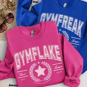 May include: Two sweatshirts, one royal blue and one hot pink, are displayed. The blue sweatshirt has the text "GYMFREAK" in white, while the pink one says "GYMFLAKE". Both have additional text and a star graphic.