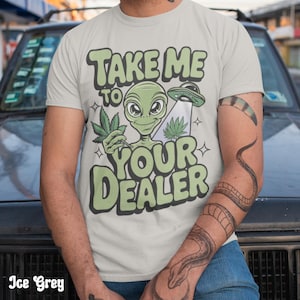 Op de afbeelding: Ijsgrijs t-shirt met een groene alien die een cannabisblad en een UFO vasthoudt. De tekst "TAKE ME TO YOUR DEALER" is groen. Het shirt is gemaakt van een zacht materiaal. Er is ook een slangentattoo te zien.
