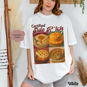May include: White t-shirt with the text "Certified Side B*tch" in brown and red script. The shirt features a graphic of four different side dishes in bowls, including mashed potatoes, mac and cheese, and green bean casserole.