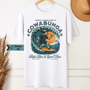 May include: White unisex t-shirt featuring a cartoon cow surfing a wave with the text "COWABUNGA" and "High Tides & Good Vibes". The design includes a sun, stars, and the phrase "LIFE'S A WAVE-RIDE IT".