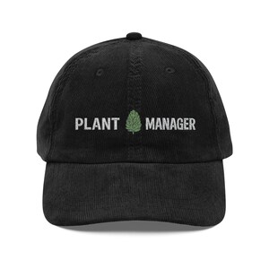 May include: Black corduroy baseball cap with the text "PLANT MANAGER" in white, with a green plant graphic in the center. The cap has a curved brim and a button on top. A casual hat for plant enthusiasts.