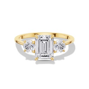May include: A gold ring featuring a large, rectangular-cut diamond center stone flanked by two smaller, round-cut diamonds. The ring has a classic three-stone design, with a polished gold band. The diamonds sparkle brightly against a white background.