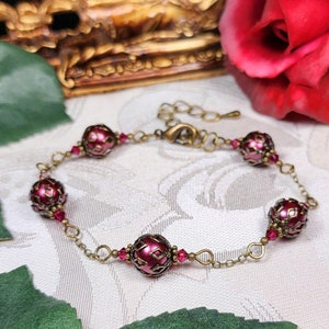 May include: A burgundy and gold bracelet with a chain and five round beads. The beads have a patterned design and are accented with small red crystals.
