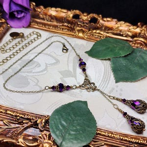 May include: A purple and gold necklace with a delicate chain and two dangling teardrop-shaped charms. The charms are adorned with intricate filigree designs and feature a purple iridescent finish. The necklace is displayed on a gold-colored surface with green leaves.