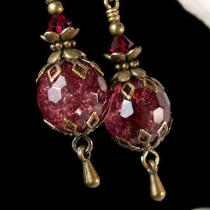 May include: A pair of dangle earrings featuring faceted, deep red beads encased in ornate, antique gold-toned metalwork. The earrings have a vintage aesthetic, with small red crystal accents and teardrop-shaped dangles.
