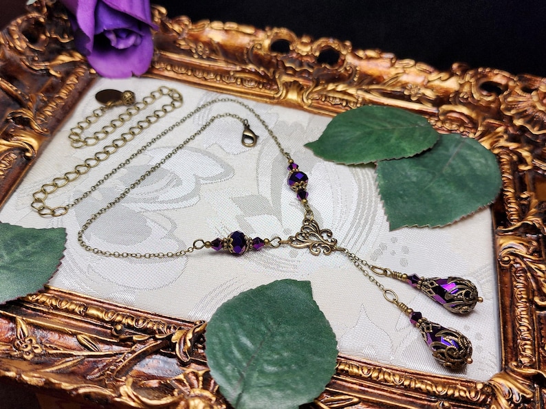 May include: A purple and gold necklace with a delicate chain and two dangling teardrop-shaped charms. The charms are adorned with intricate filigree designs and feature a purple iridescent finish. The necklace is displayed on a gold-colored surface with green leaves.