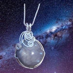 May include: A silver pendant necklace featuring a large, round, gray gemstone. The gemstone is encased in a silver wire frame with decorative swirls. The necklace hangs against a backdrop of a starry night sky.