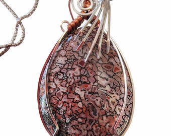 May include: A teardrop-shaped pendant with a patterned stone in shades of red, black, and white, set in a silver-tone metal frame. The pendant has a silver chain and copper accents.