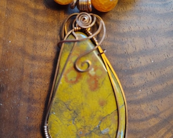 May include: A handmade necklace with a large, teardrop-shaped pendant made from a yellow and green stone, encased in copper wire. The necklace has a strand of orange and yellow beads, creating a unique and earthy jewellery piece.