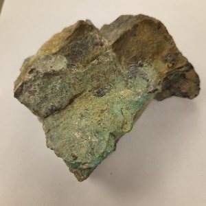 May include: A raw, unpolished mineral specimen featuring a rough, jagged texture. The stone displays a mix of earthy brown and tan tones across its upper sections, transitioning into a vibrant, textured patch of light turquoise and mint green.