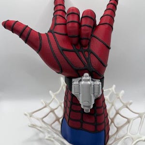 May include: A red and blue hand sculpture featuring a black web pattern and a silver wrist-mounted device. The hand is posed in a gesture with the thumb, index, and pinky fingers extended, resting on a white, web-like base structure.