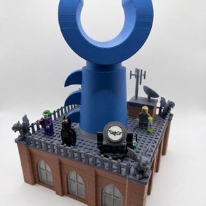 May include: A building rooftop display constructed from plastic bricks featuring a large blue C-shaped structure, a bat signal spotlight, and four gray gargoyles. Several small character figures are positioned on the roof, which has brick-patterned walls and arched windows.