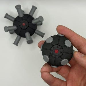 May include: Two black and gray spherical objects with cylindrical gray protrusions. One is held in a hand, the other rests on a white surface. Each sphere has a red center and gray circular accents.