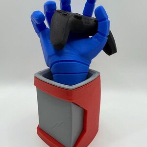 May include: A blue, 3D-printed hand holding a black game controller. The hand is mounted on a red and gray container, likely for desk organization or display. The design is modern and geometric.