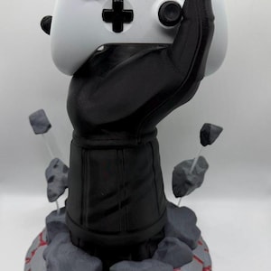 May include: A white Xbox controller is held by a black hand-shaped stand. The stand is on a gray and red stone base with gray rocks around the base. The controller has black buttons and a black cross-shaped directional pad.