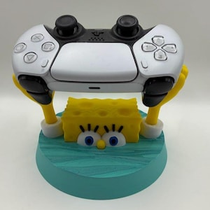 May include: A white and black video game controller is held by a yellow SpongeBob SquarePants-themed stand. The stand has a blue base with a water-like design. The controller has black joysticks and buttons.