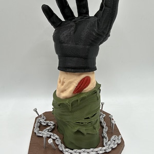 May include: A 3D printed sculpture of a black gloved hand emerging from a severed arm with a red wound. The arm is wrapped in a tattered olive green sleeve, positioned on a brown hexagonal base with silver chains and metal nails protruding.
