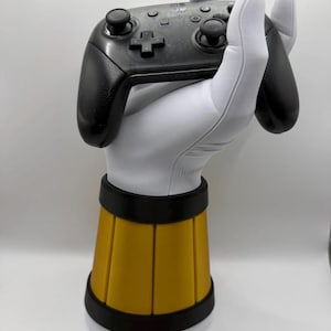 May include: A black Switch Pro controller held by a white gloved hand, mounted on a yellow and black base. The controller has black thumbsticks and buttons. The base is cylindrical with a yellow center and black trim.