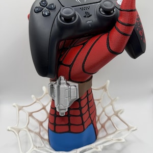 May include: A red and blue textured hand sculpture with black webbing details and a silver wrist cuff, holding a black wireless game controller. The controller features a white spider logo on the touchpad and a white web pattern on the right handle.
